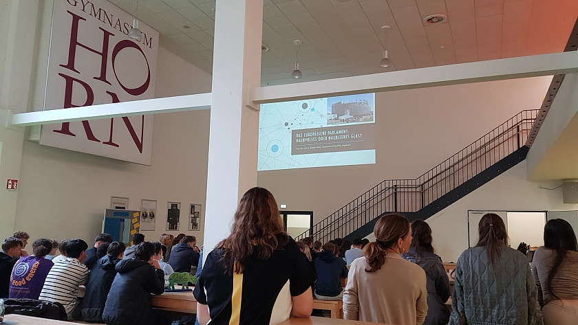 Foto des Vortrags in der Mensa der Schule mit Rücken von Schülern und weiter hinten der Vortrag an der Wand per Beamer projiziert