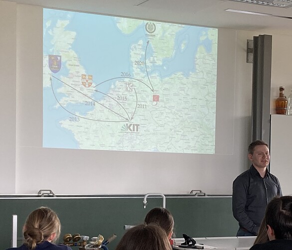 Foto des Vortrags in der Mensa der Schule mit Rücken von Schülern und weiter hinten der Vortrag an der Wand per Beamer projiziert. Der Redner steht davor.