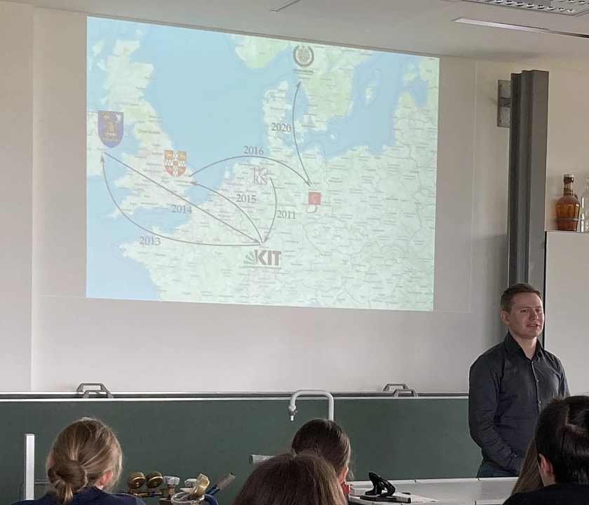Foto des Vortrags in der Mensa der Schule mit Rücken von Schülern und weiter hinten der Vortrag an der Wand per Beamer projiziert. Der Redner steht davor.