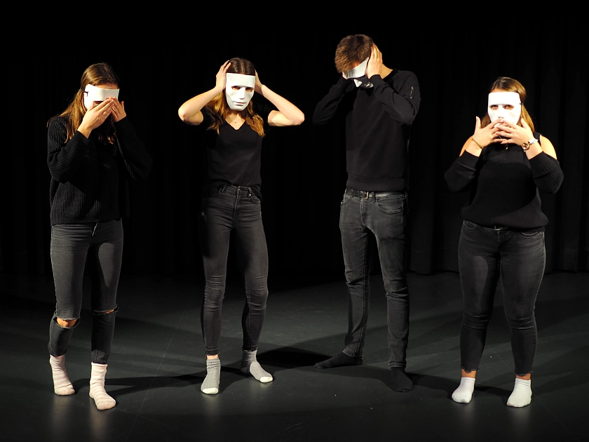 Foto von 4 Schülern in schwarz mit weißen MAsken auf der Bühne