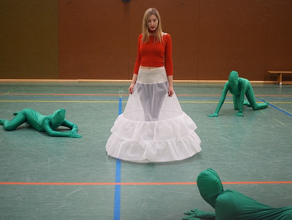 Foto der Performance mit einer Frau und Kleid in der Mitte und 3 grünen Menschen um sie herum auf dem Boden