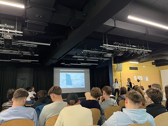 Foto des Vortrags in der Mensa der Schule mit Rücken von Schülern und weiter hinten der Vortrag an der Wand per Beamer projiziert